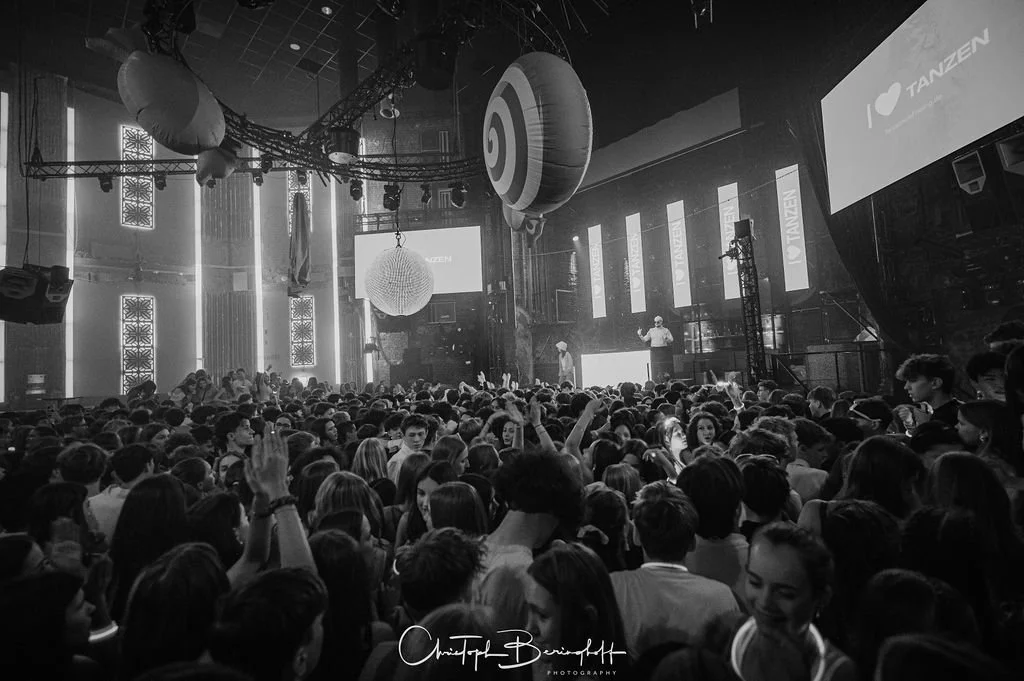 A large crowd is dancing and celebrating in a nightclub filled with colorful lights and a stage where someone is speaking. There are large LED screens and balloons hanging from the ceiling.