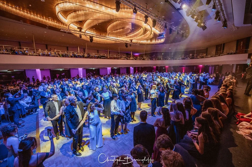 Bilder von einer Tanzveranstaltung in einem großen Saal mit vielen tanzenden Paaren, Zuschauern am Rand und buntem Licht, das den Raum beleuchtet.