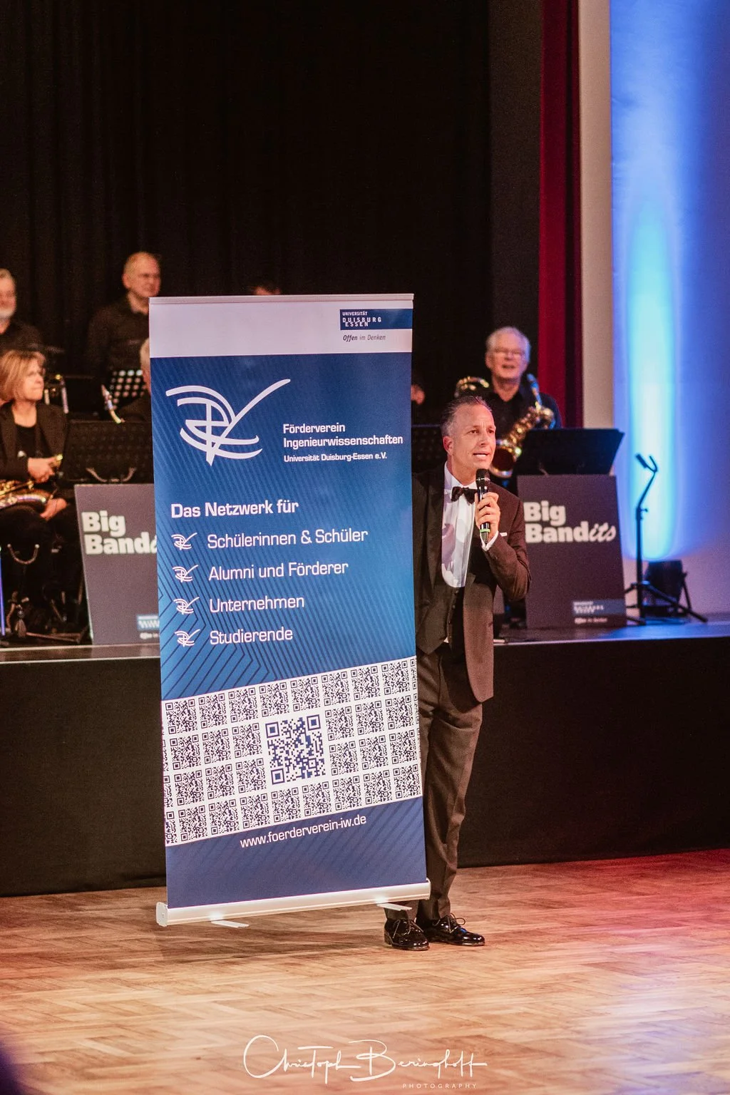 Ein Mann in Smoking hält in einem Saal ein Mikrofon und steht neben einem blauen Banner für den Förderverein Ingenieurwissenschaften der Universität Duisburg-Essen. Im Hintergrund spielen Musiker auf einer Bühne.