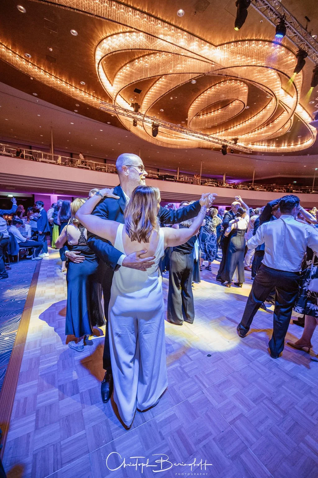 Menschen tanzen auf einer eleganten Feier in einem großen Saal mit einer beeindruckenden, geschwungenen, goldenen Deckenbeleuchtung.