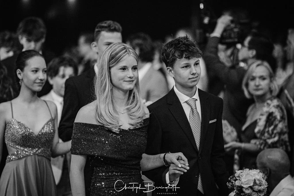 Junge Menschen bei einer formellen Veranstaltung, einer trägt ein schwarzes Glitzerkleid, die anderen tragen Anzüge und Kleider, einige halten Blumen, im Hintergrund sind weitere Personen und eine Kamera.