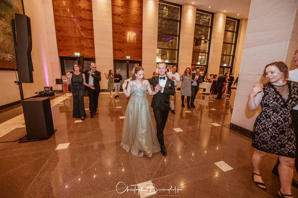 Viele Gäste tanzen und feiern auf einer Hochzeitsfeier in einem eleganten Ballsaal mit hohen Fenstern und Holzdekorationen.