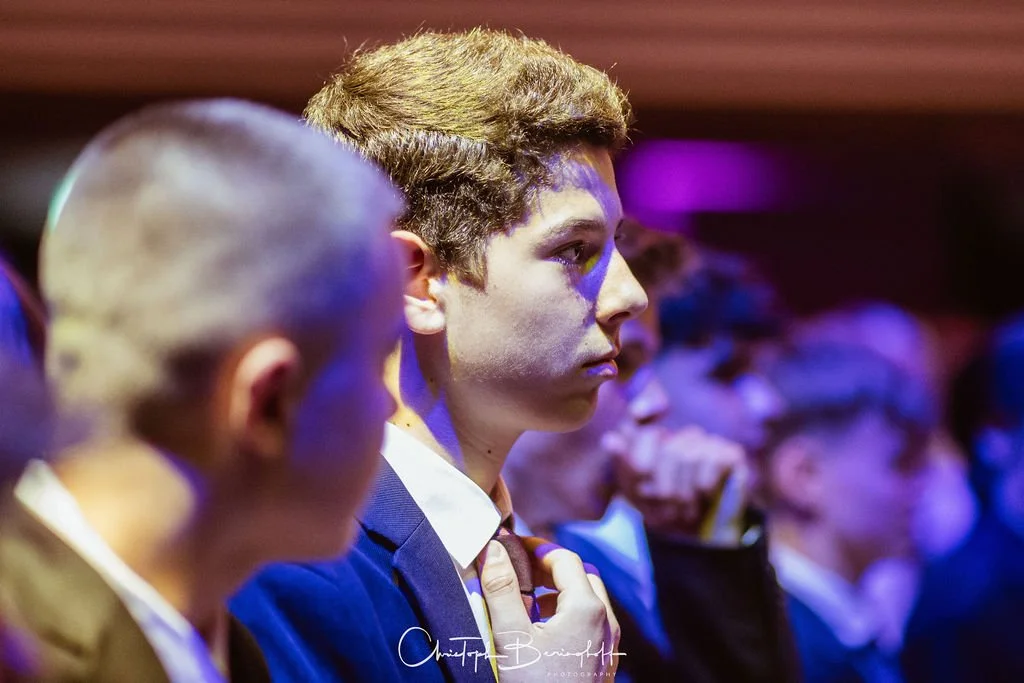 Junge Mann und weitere Personen bei einer Veranstaltung, Fokus auf einen Jungen im Profil, helle Haare, formelle Kleidung, im Hintergrund ebenfalls mehr Menschen.