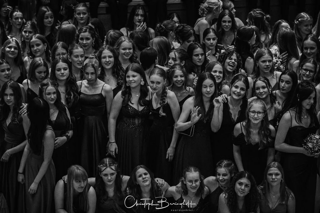 Viele junge Frauen in schwarzen Kleidern bei einer Feier, einige lachen und winken, im Hintergrund eine dunkle Wand.