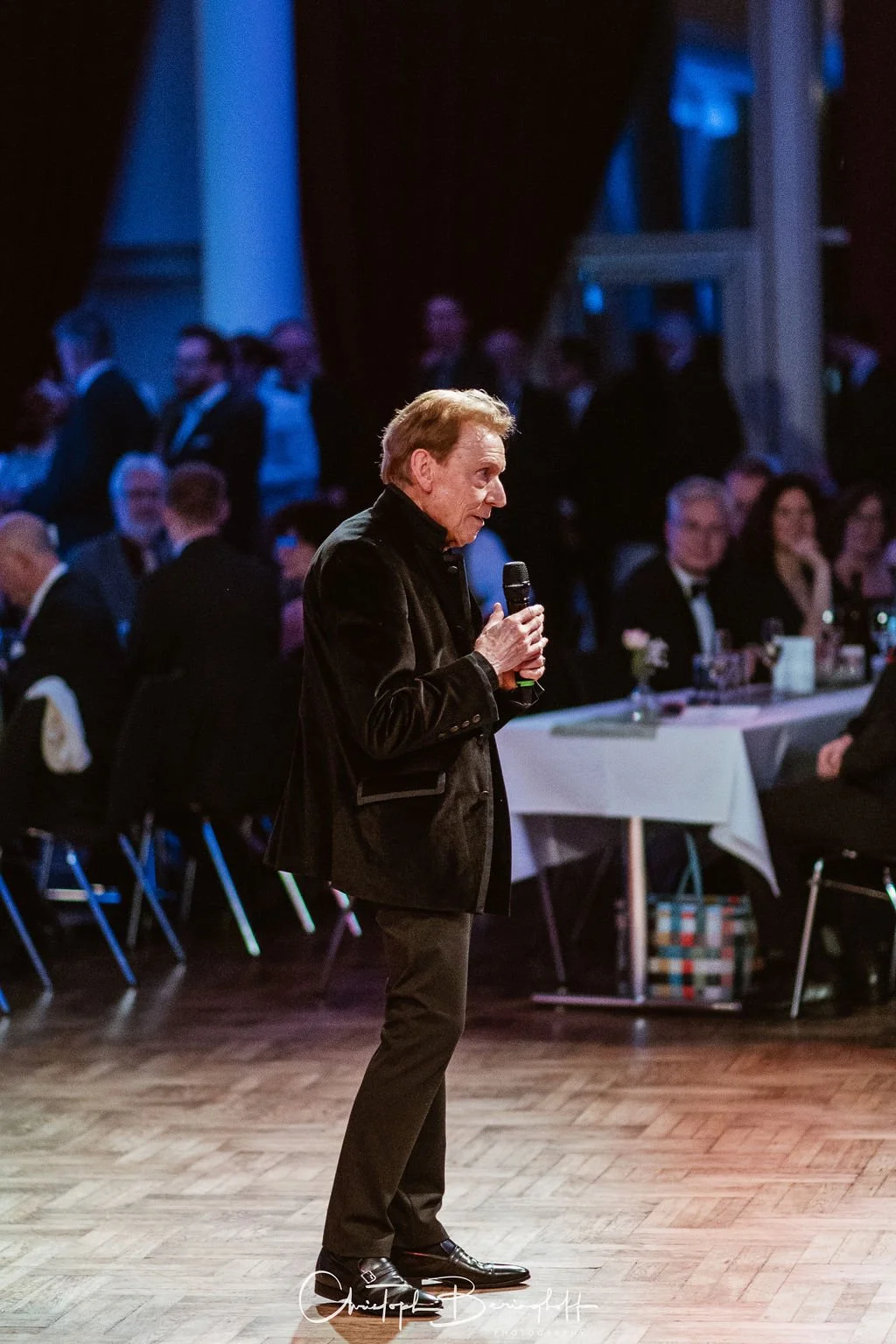 Ein älterer Mann steht auf einer Bühne, hält ein Mikrofon in der Hand und spricht vor einer vollbesetzten Tafel in einem dunklen Raum bei einer formellen Veranstaltung.