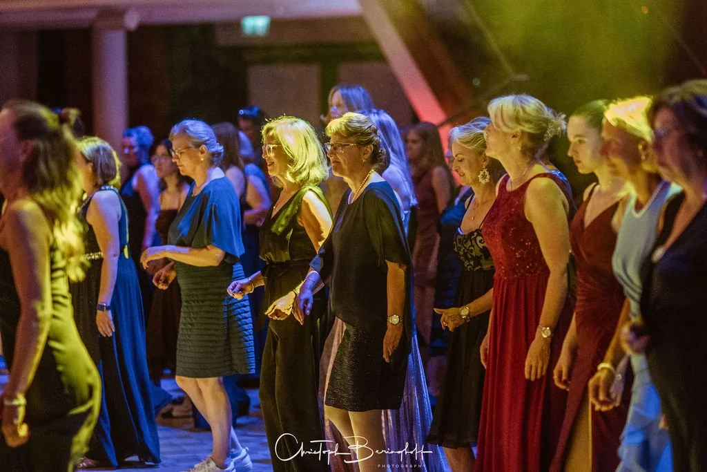 Menschen tanzen bei einer Tanzveranstaltung, überwiegend Frauen in eleganten Abendkleidern, in einem festlich beleuchteten Raum.