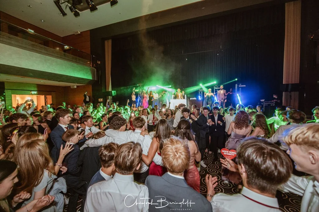 Tanzveranstaltung mit vielen Jugendlichen auf der Tanzfläche, eine Band auf der Bühne spielt, bunte Lichteffekte, im Hintergrund eine Bar, Party mit lebhafter Atmosphäre.