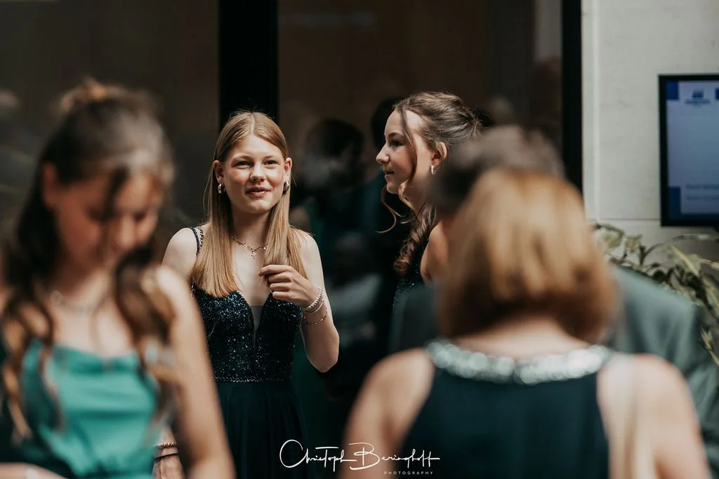 Gruppe junger Frauen bei einer Feier, unterhalten sich in einem Raum mit dunklem Hintergrund und einem Bildschirm auf der rechten Seite.