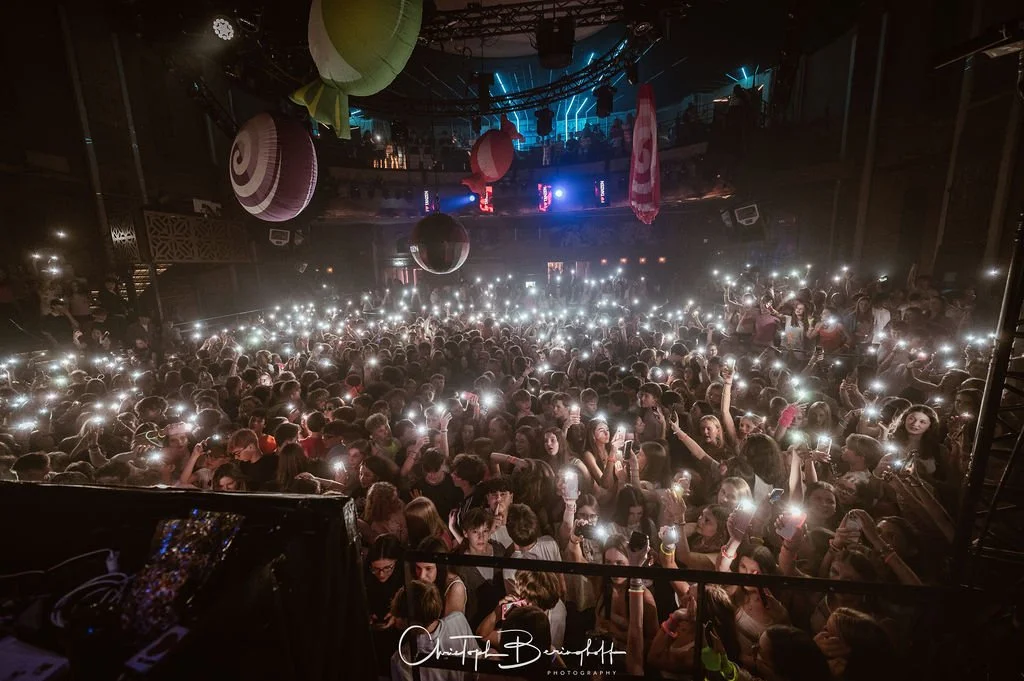 A crowd at a concert; many people are holding cell phones with the flashlights on; colorful speakers hang from the ceiling; a dark atmosphere with flashes of light.