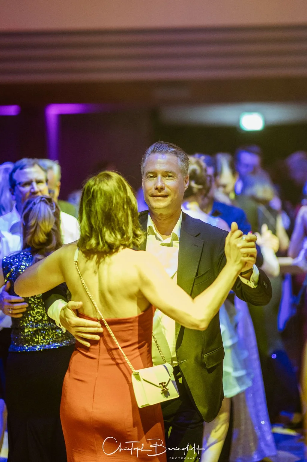 Ein Mann und eine Frau tanzen auf einer Party, im Hintergrund sind mehrere andere Paare, bunte Lichtverhältnisse, formelle Kleidung.