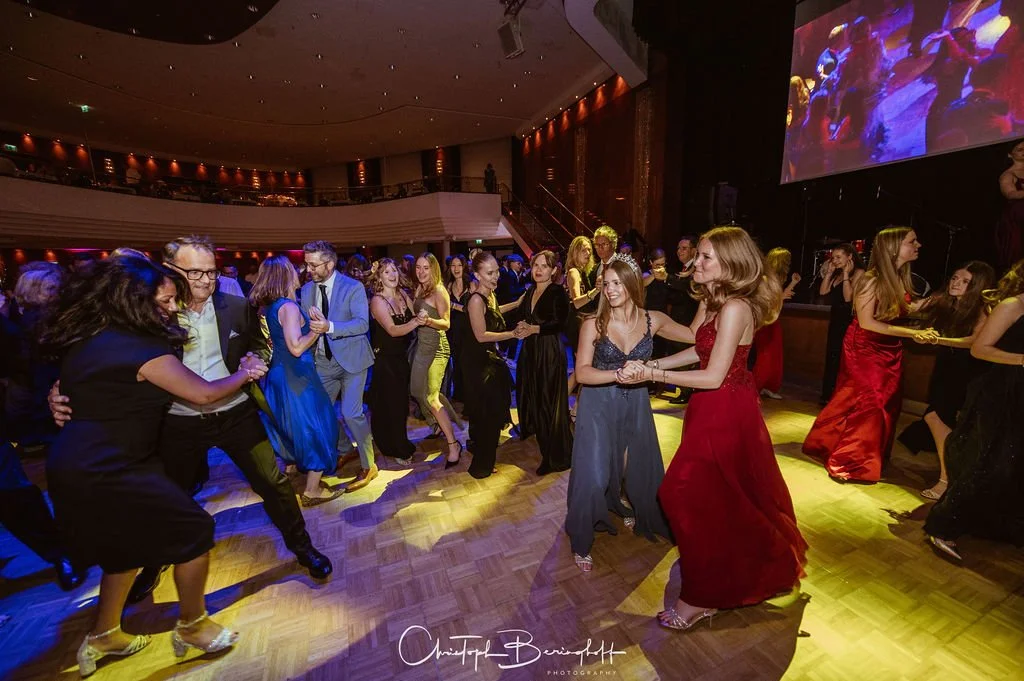 Menschen tanzen auf einer Tanzfläche bei einer Party oder einem Ball, im Hintergrund ein großes Bildschirm mit bunten Lichteffekten.