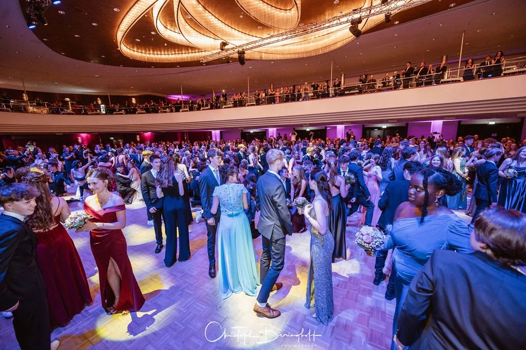 Tanzsaal mit vielen jungen Menschen, die auf einer Party oder Ball tanzen, in eleganten Kleidern und Anzügen, mit einer oberen Empore und einer modernen, gut beleuchteten Decke.
