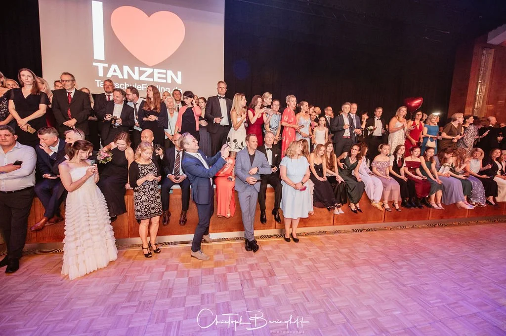 Viele Leute sitzen und stehen auf einer Bühne bei einer Veranstaltung, mit einem großen Bildschirm im Hintergrund, auf dem steht "I ❤️ TANZEN".