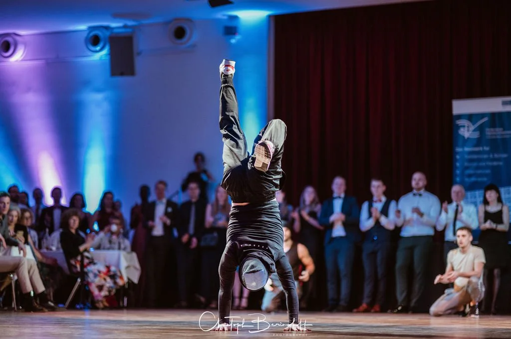 Ein Tänzer zeigt einen Handstand auf einer Bühne, während ein Publikum im Hintergrund zuschaut.