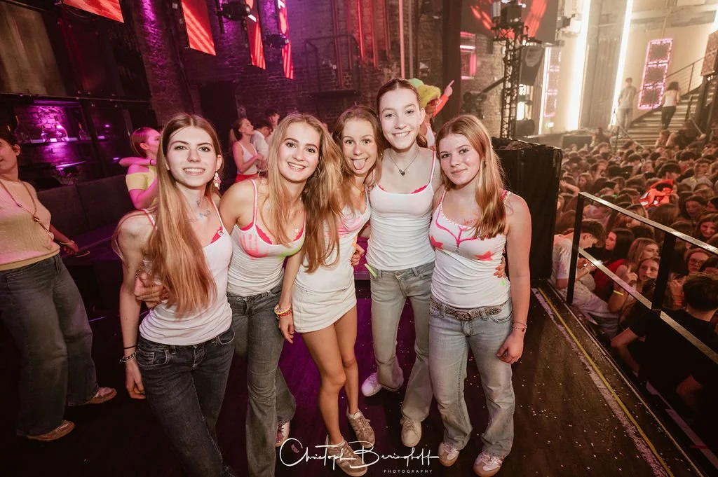 Five young women are posing in a nightclub setting with colorful lights and an audience in the background.