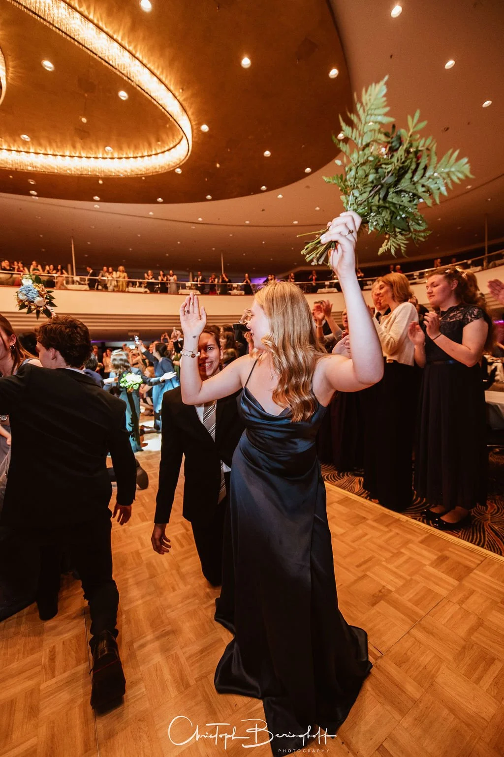 Frau in einem schwarzen Abendkleid hält eine große Blumenstrauß hoch in einem festlich dekorierten Saal, umgeben von applaudierenden Menschen.