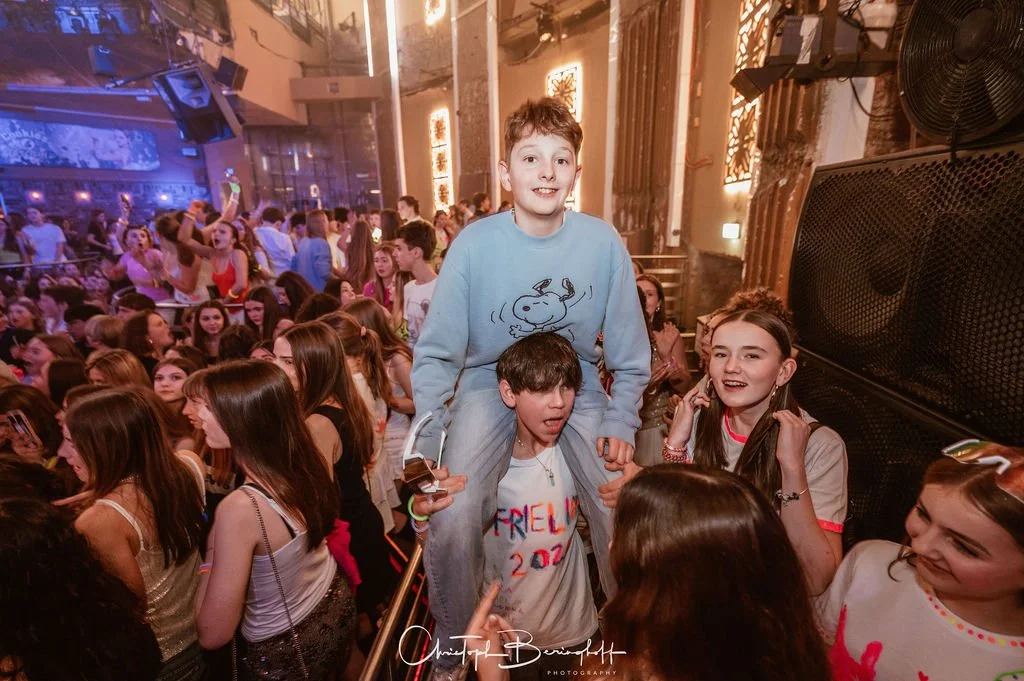 A boy is sitting on a friend's shoulders at a party with lots of teenagers who are happily celebrating in a festively decorated room.