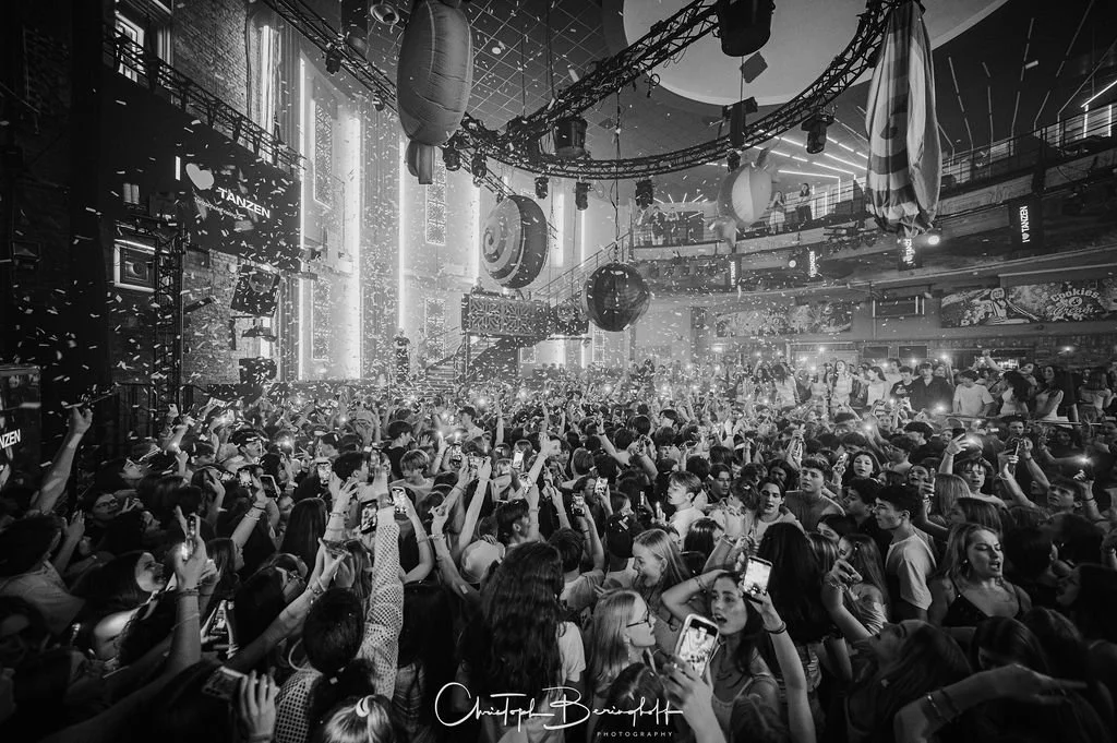 People are dancing in a dark club, surrounded by beams of light, balloons, and confetti, celebrating at a party.