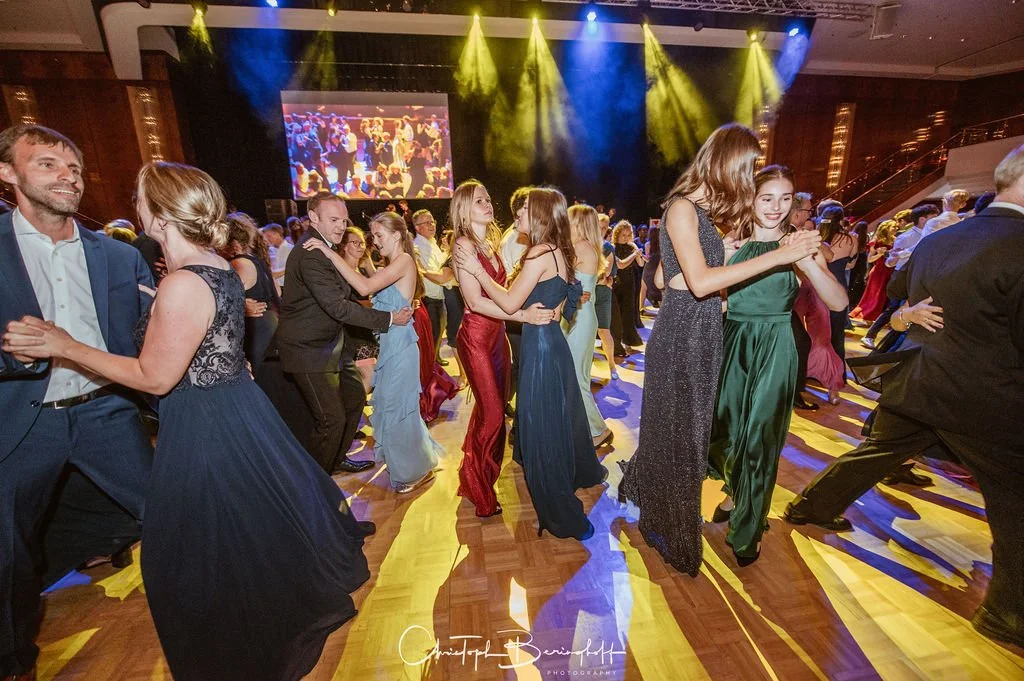 Tanzende Menschen in festlicher Kleidung bei einer Party im Ballsaal mit buntem Licht und einer großen Leinwand im Hintergrund.