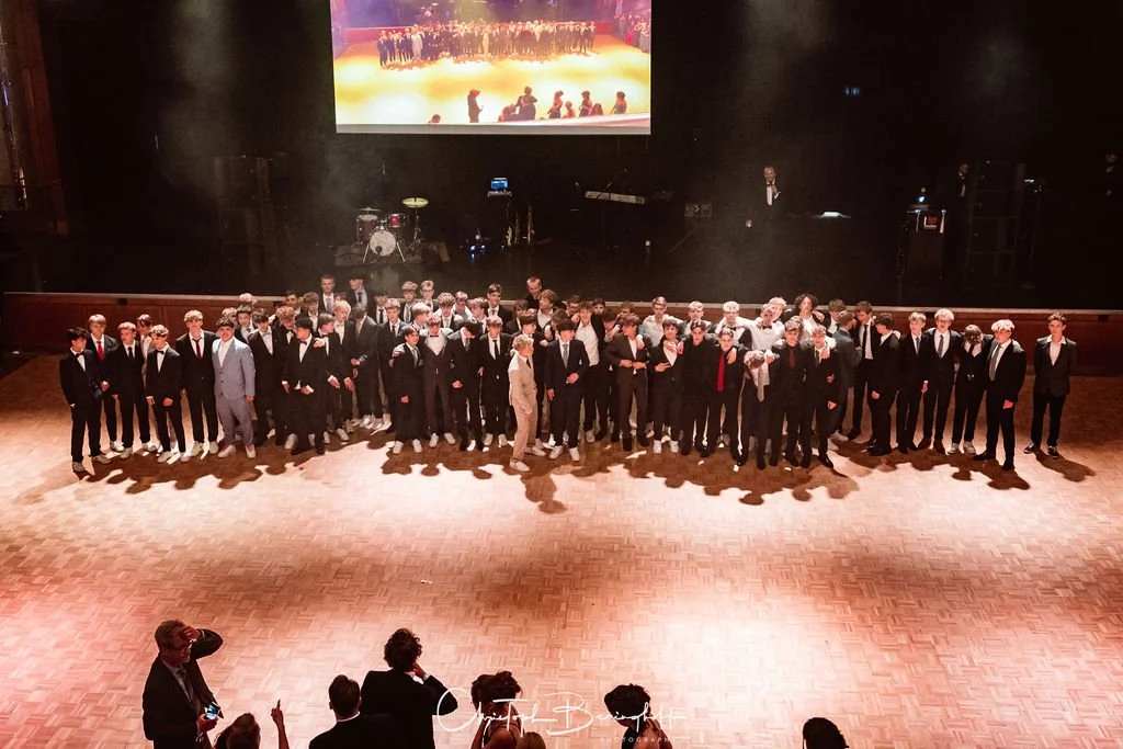 Gruppe von Jugendlichen in eleganter Kleidung auf einer Bühne vor einem großen Bildschirm, vermutlich bei einer Veranstaltung oder Gala.