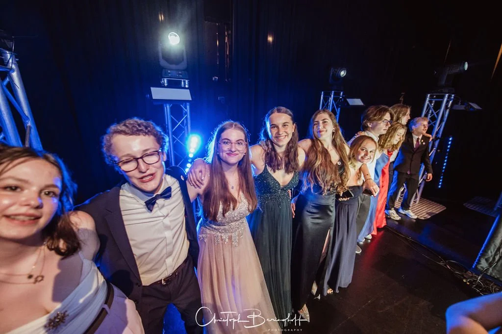Gruppe junger Menschen auf einer Bühne bei einem festlichen Anlass, alle in eleganter Kleidung, applaudierend und lachend.