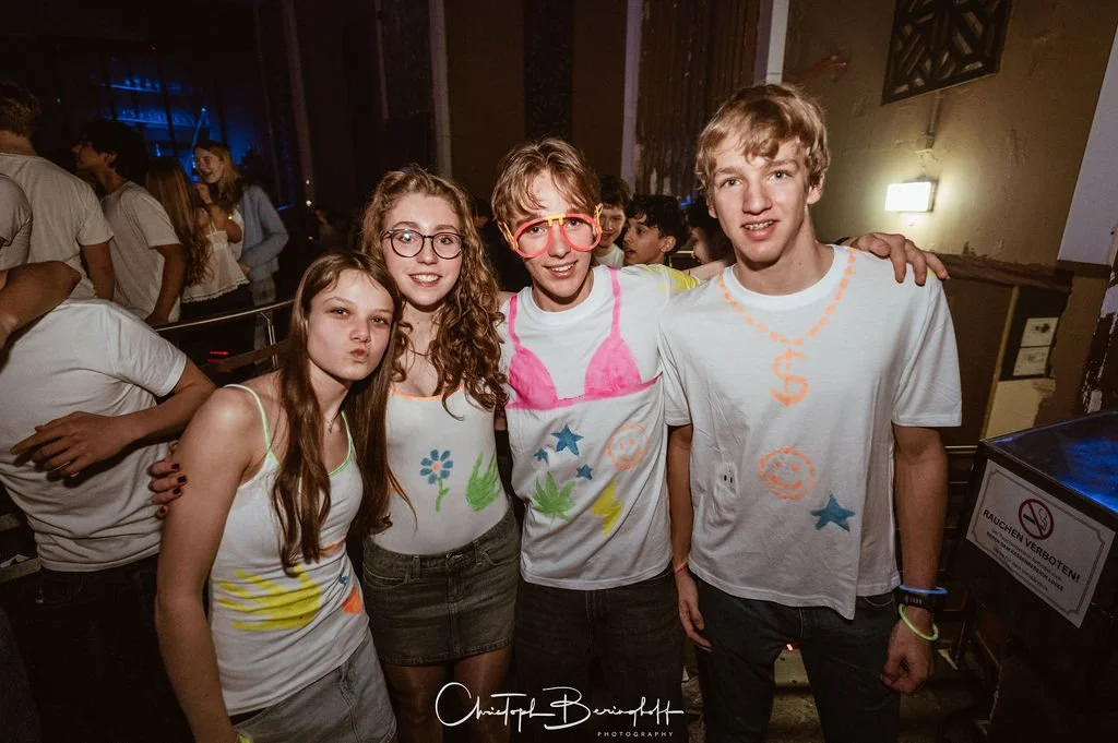 Four young people are happily posing at a party; they're all wearing hand-painted T-shirts with colorful designs.