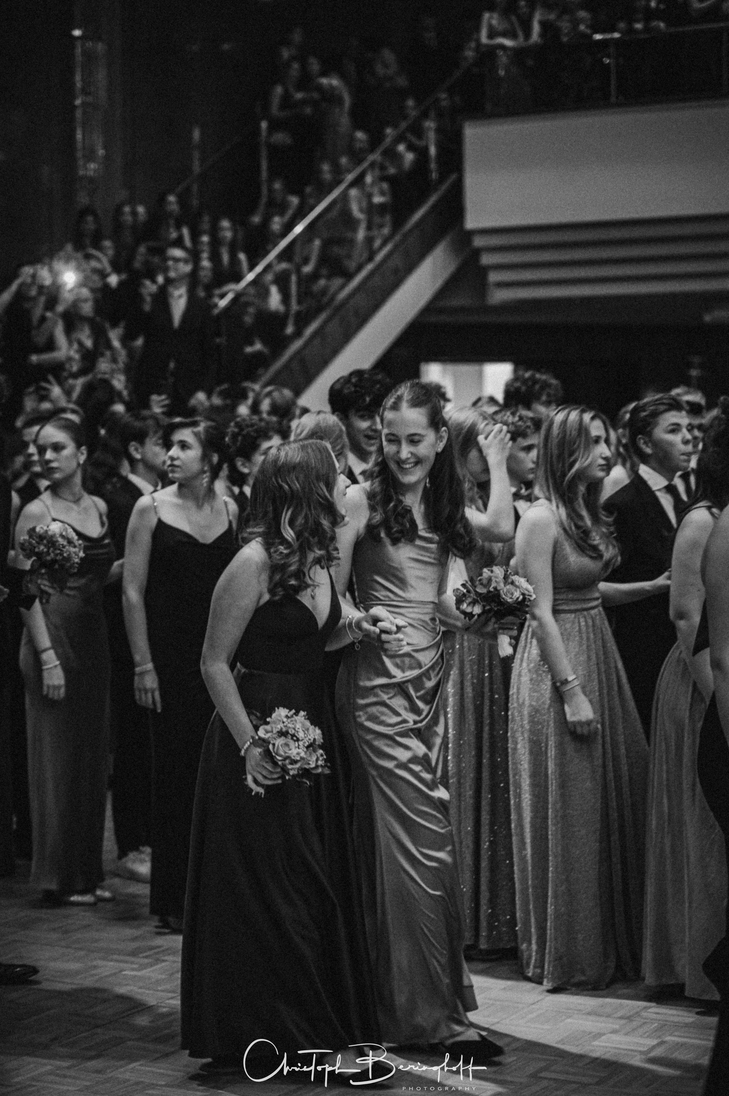 Menschenmenge bei einer formellen Veranstaltung, hauptsächlich junge Frauen in eleganten Kleidern, einige halten Blumen, im Hintergrund Treppen und Zuschauer, schwarz-weiß Fotografie.