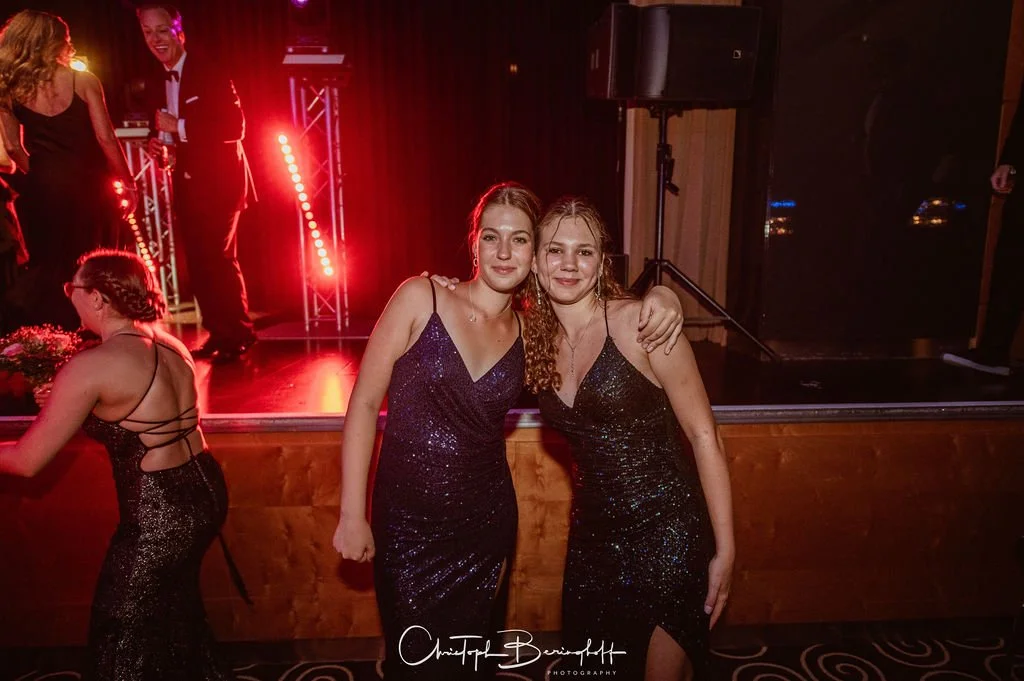 Zwei junge Frauen in dunklen, glitzernden Kleidern posieren auf einer Party vor einer Bühne mit roter Beleuchtung.