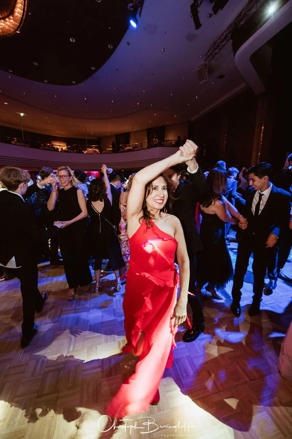 Menschen tanzen auf einer Party im eleganten Stil in einem großen Saal mit bunten Lichtern, eine Frau in rotem Kleid im Vordergrund tanzt fröhlich.