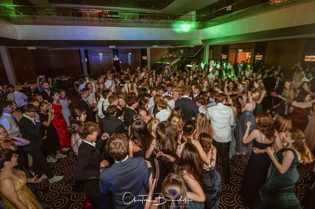Eine große Gruppe junger Menschen tanzt auf einer Party in einem eleganten Veranstaltungsraum mit dunklen Wänden, einer Holztreppe und grüner Beleuchtung.