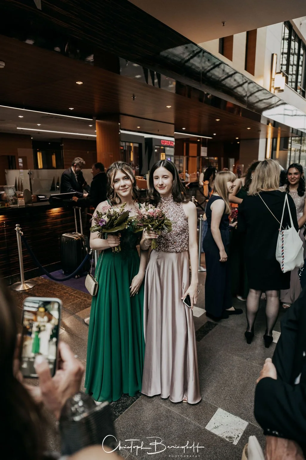 Zwei junge Frauen in eleganten Abendkleidern, die Blumen halten, posieren im Eingangsbereich eines Hotels oder Veranstaltungsortes bei einer formellen Veranstaltung.