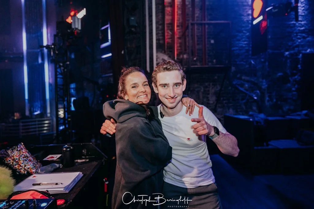 Two people are hugging each other in a dark room lit by neon lights, smiling.