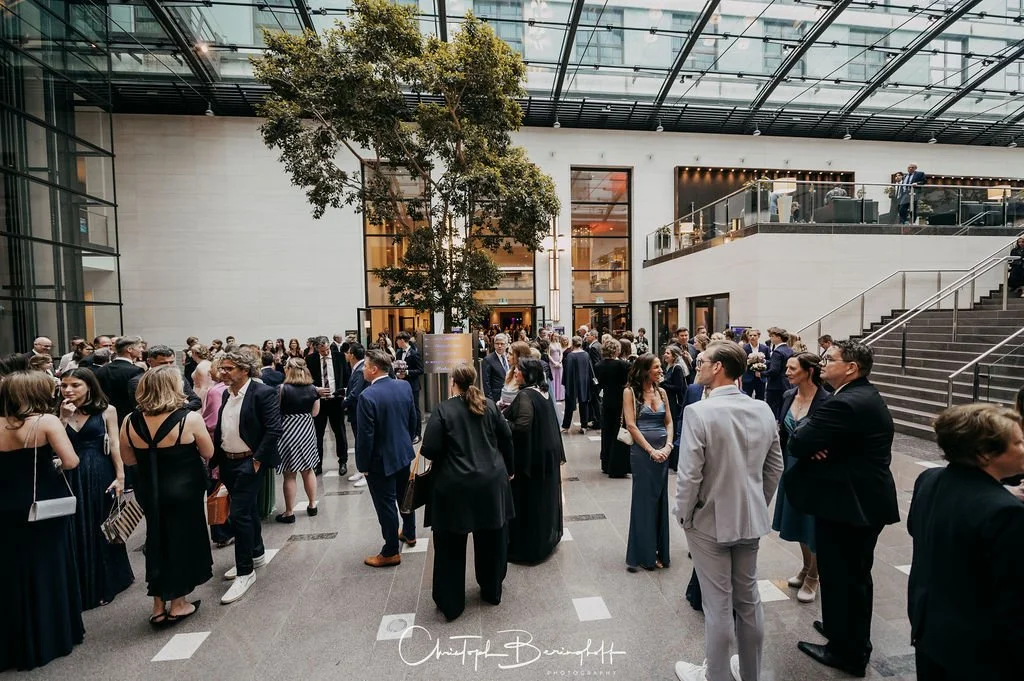 Menschen in eleganter Kleidung bei einer Netzwerkveranstaltung in einem modernen, hellen Atrium mit Glasdecke.
