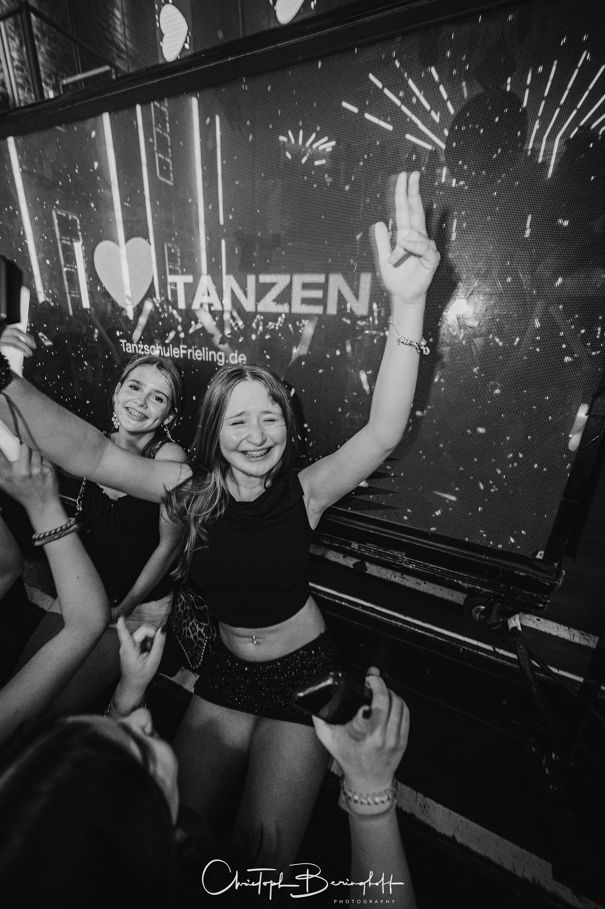 Zwei lachende junge Mädchen feiern in einem Club, eine hält ein Smartphone und posiert vor einer LED-Wand mit der Aufschrift "Ich liebe Tanzen".