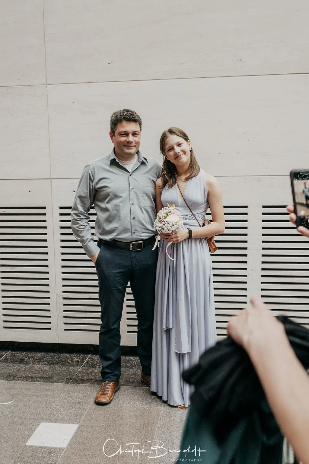 Ein Mann und eine junge Frau stehen nebeneinander vor einer hellen Wand, der Mann trägt ein Hemd und dunkle Hose, die Frau trägt ein elegantes Kleid und hält einen Blumenstrauß. Es sieht nach einer Feier aus, Kamera wird von einer Person rechts gehal