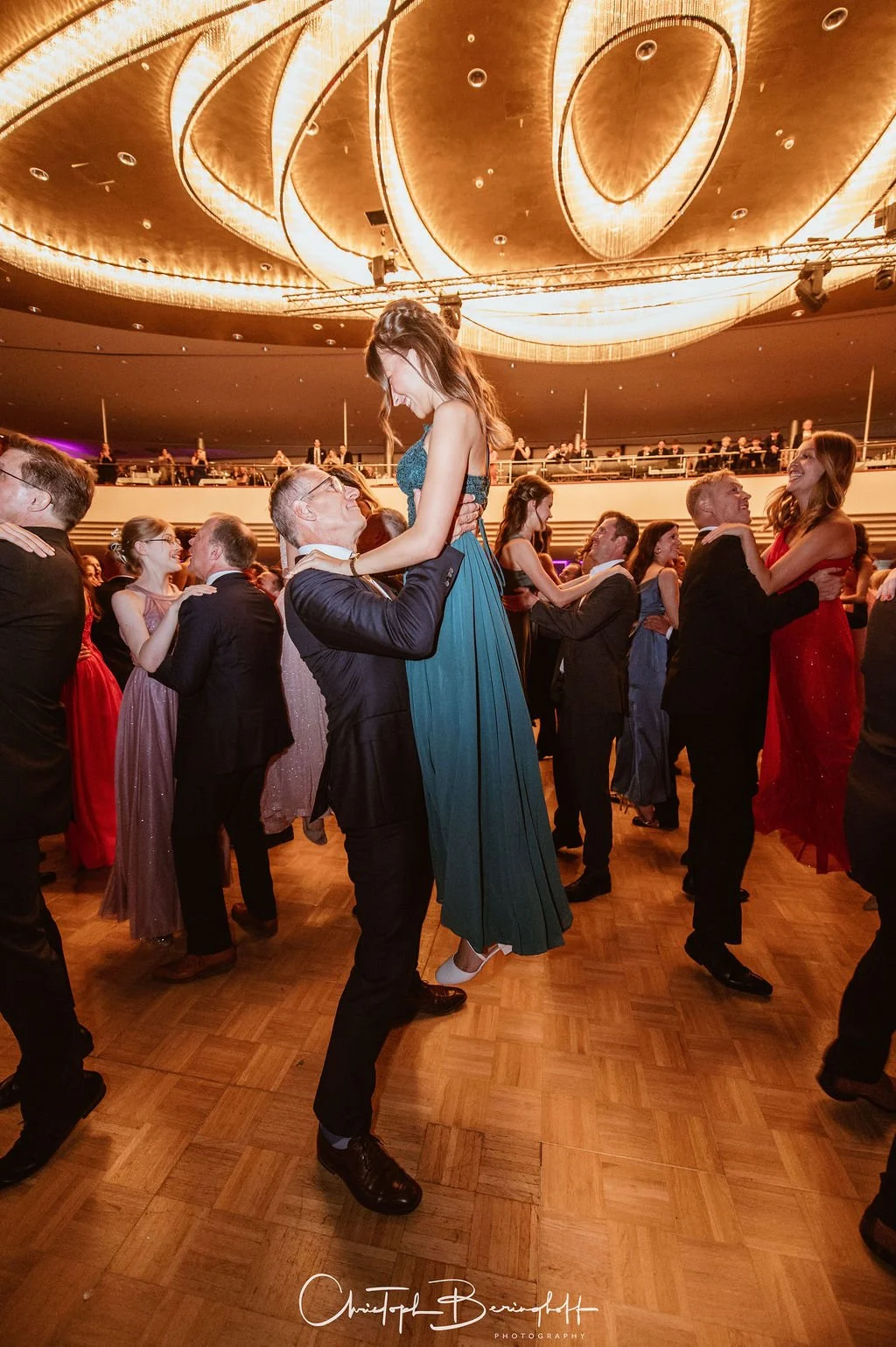 Paare tanzen bei einer formellen Veranstaltung auf einer Tanzfläche im Ballsaal, während die Decke kunstvoll beleuchtet ist.