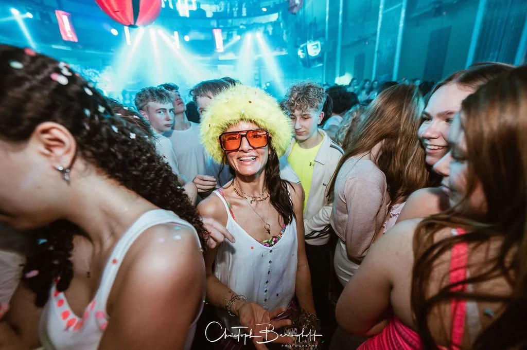 People are celebrating at a party with colorful lights and confetti; one woman is wearing a striking yellow hat and large sunglasses.