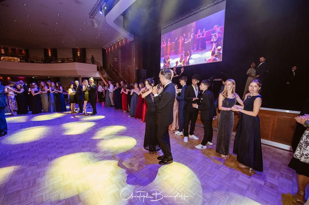 Menschen tanzen bei einer formellen Veranstaltung in einem eleganten Saal, mit einer großen Leinwand im Hintergrund, die eine Live-Performance zeigt.