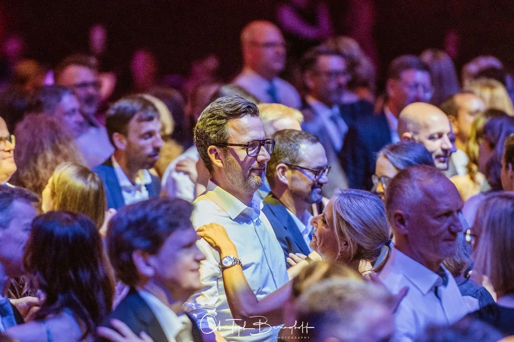 Menschen beim festlichen Event, lachend und im Gespräch, in eleganter Kleidung, mit buntem Licht im Hintergrund.
