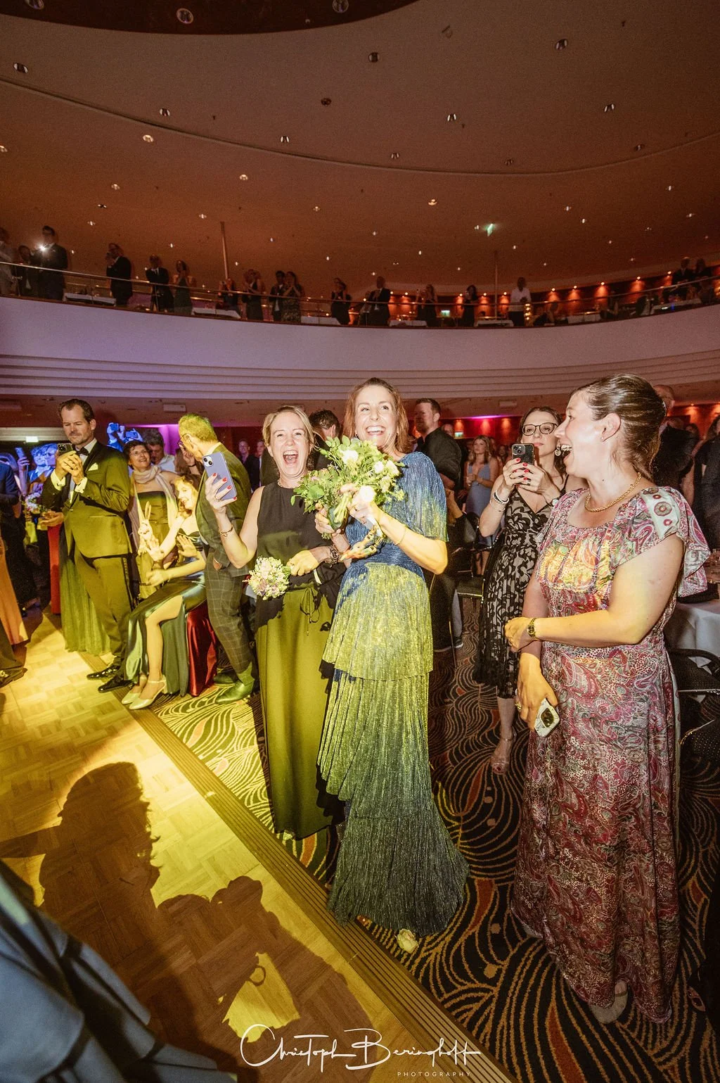 Menschen auf einer festlichen Veranstaltung, lachen und feiern. Zwei Frauen im Mittelpunkt halten einen Blumenstrauß, umrahmt von weiteren Gästen in eleganter Kleidung in einem großen, gut beleuchteten Saal.