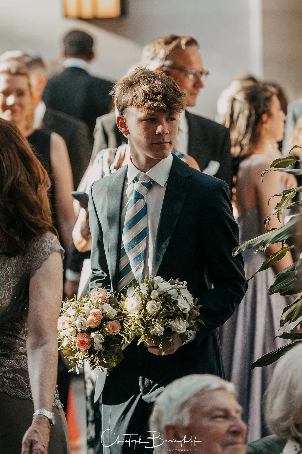 Junger Mann in Anzug mit Blumenstrauß bei einer formellen Veranstaltung, vermutlich einer Hochzeitsfeier, umgeben von anderen Gästen in eleganter Kleidung.