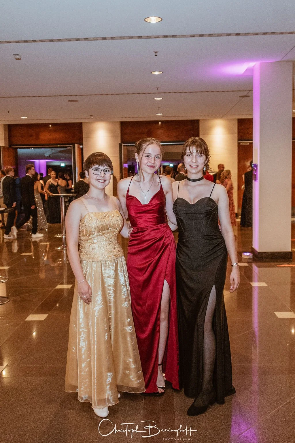 Drei junge Frauen in eleganten Abendkleidern posieren in einem festlich dekorierten Raum, vermutlich auf einer Gala oder Ball.