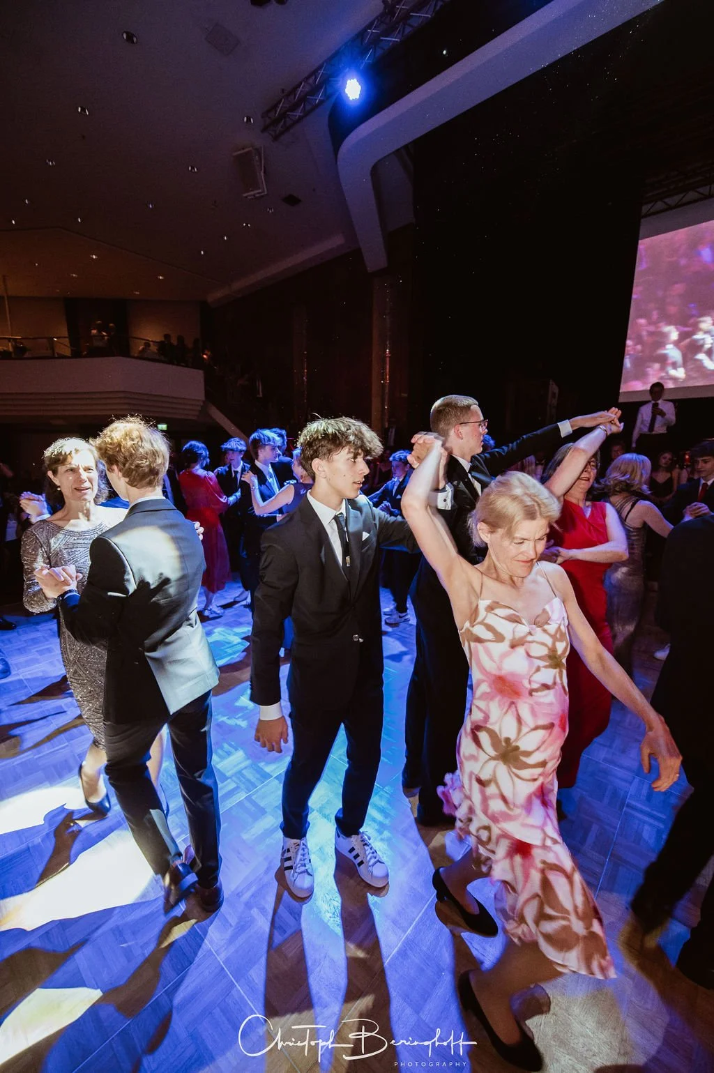 Menschen tanzen auf einer Party im Festsaal, viele in eleganter Kleidung, bunte Lichter und eine große Leinwand im Hintergrund.