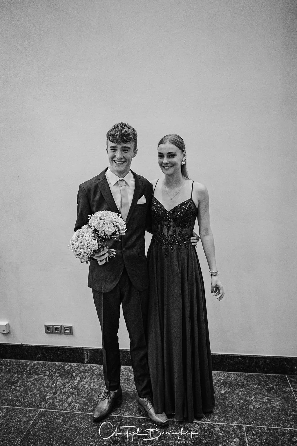 Junge Frau und junger Mann in formaler Kleidung, der Mann hält einen Blumenstrauß, stehen nebeneinander vor einer einfachen Wand, beide lachen und schauen freundlich in die Kamera.