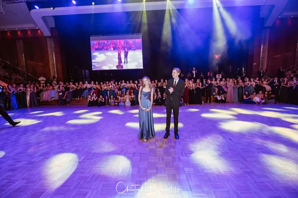 Ein festliches Ballsaal mit Tanzpaar in der Mitte, umgeben von zahlreichen Gästen in formeller Kleidung. Es gibt eine große Leinwand im Hintergrund und bunte Scheinwerfer auf dem Parkett.