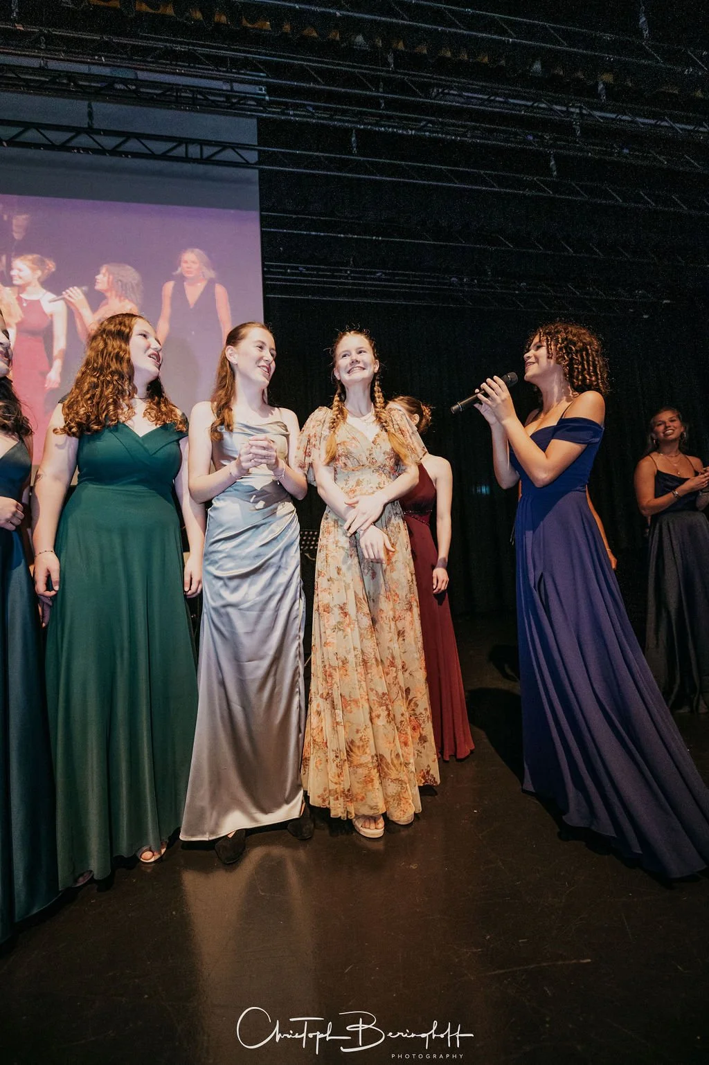 Mehrere Frauen auf einer Bühne bei einer Preisverleihung, einige tragen elegante lange Kleider, eine Frau mit dunklem, lockigem Haar hält ein Mikrofon und spricht zu einer Gruppe von Frauen.