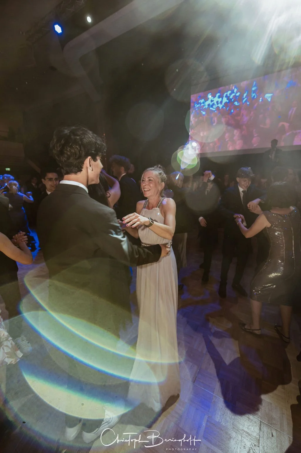 Menschen tanzen auf einer Party, eine Frau im weißen Kleid und ein Mann in einem schwarzen Anzug lächeln einander an.