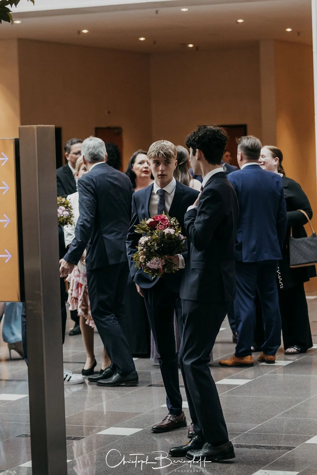 Zwei junge Männer in Anzügen tauschen sich bei einer formellen Veranstaltung aus, einer hält einen Blumenstrauß, während im Hintergrund weitere Personen in formeller Kleidung stehen und sprechen.
