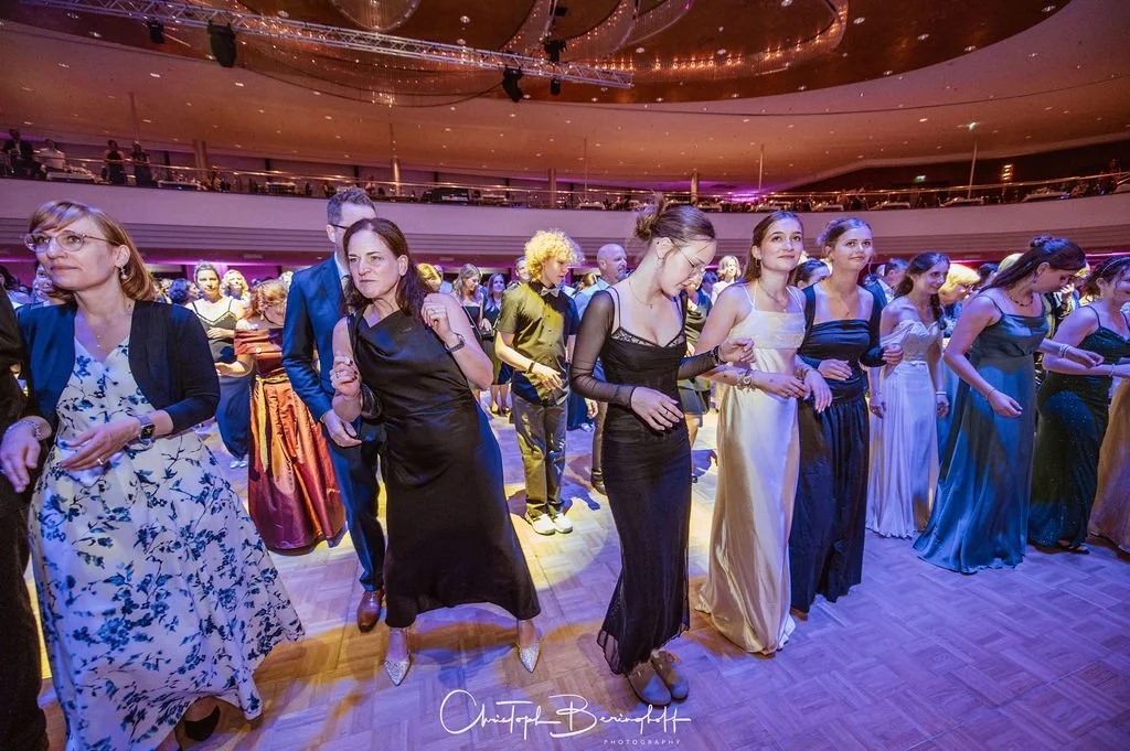 Menschen tanzen in eleganten Abendkleidern in einem festlichen Saal.