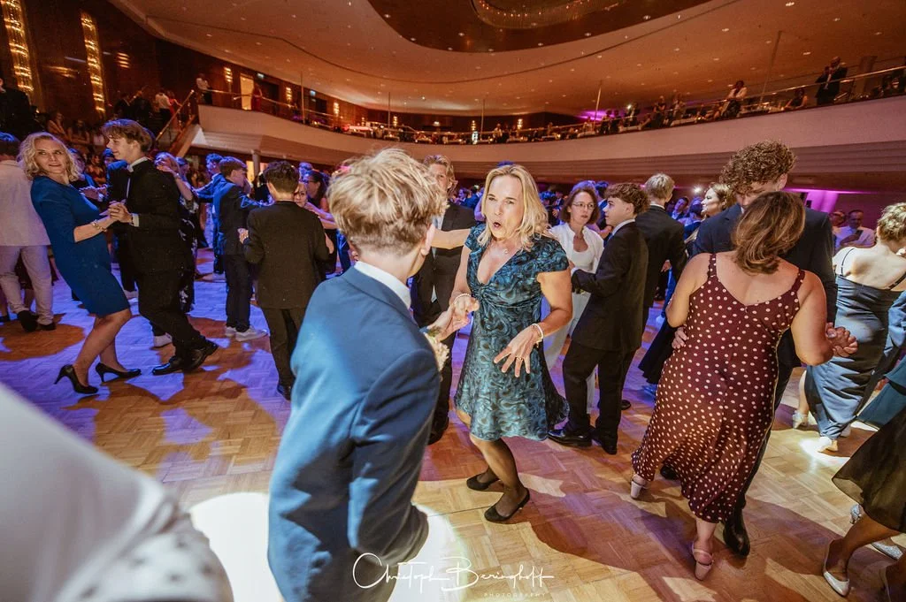 Menschen tanzen auf einer festlichen Veranstaltung in einem großen Ballsaal mit Holzboden und moderner Architektur.